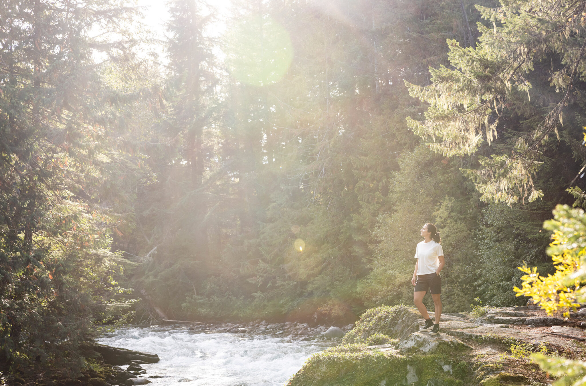 A woman stands in the sun on the bank of a river with the lush forest behind her in Whistler.
